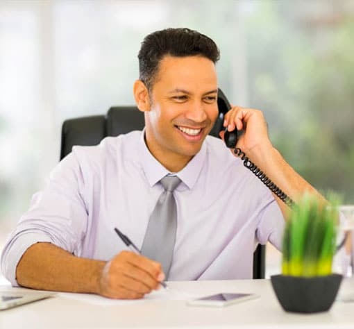 Rapaz em um escritório, falando sorridente ao telefone enquanto realiza anotações.